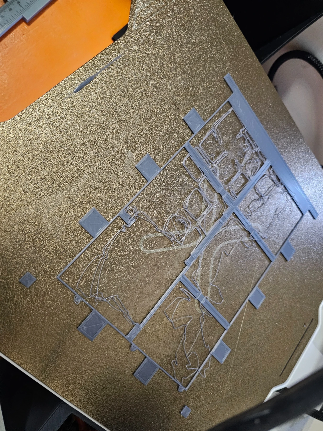 Real photograph of an unsuccessful gray floor-frame print on a textured build plate, with loose filament visible.